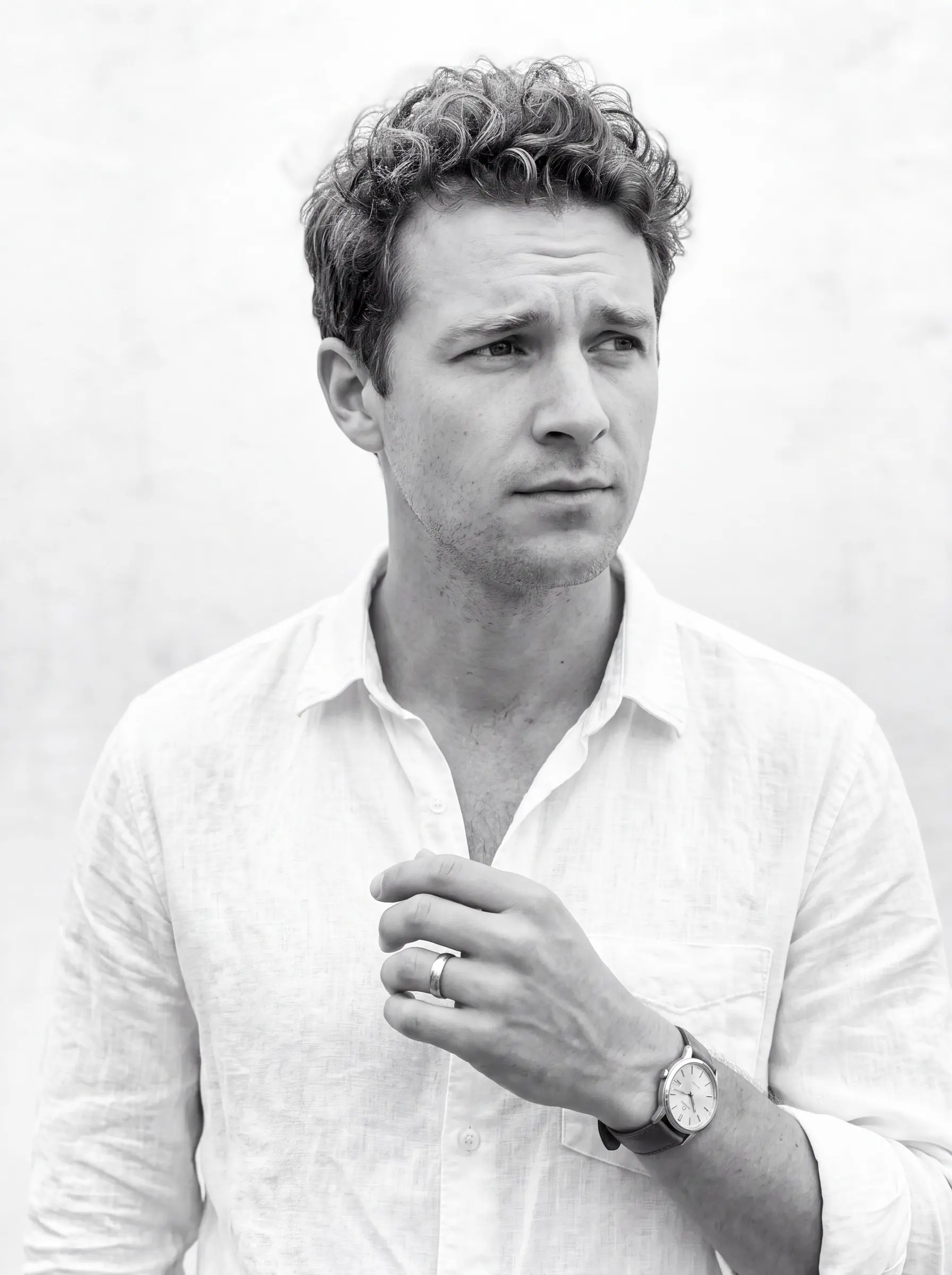 Black-and-white portrait of a man with short curly hair in an open-collar white linen shirt, looking to the side, one hand near his chest showing a ring and a watch, against a pale wall.