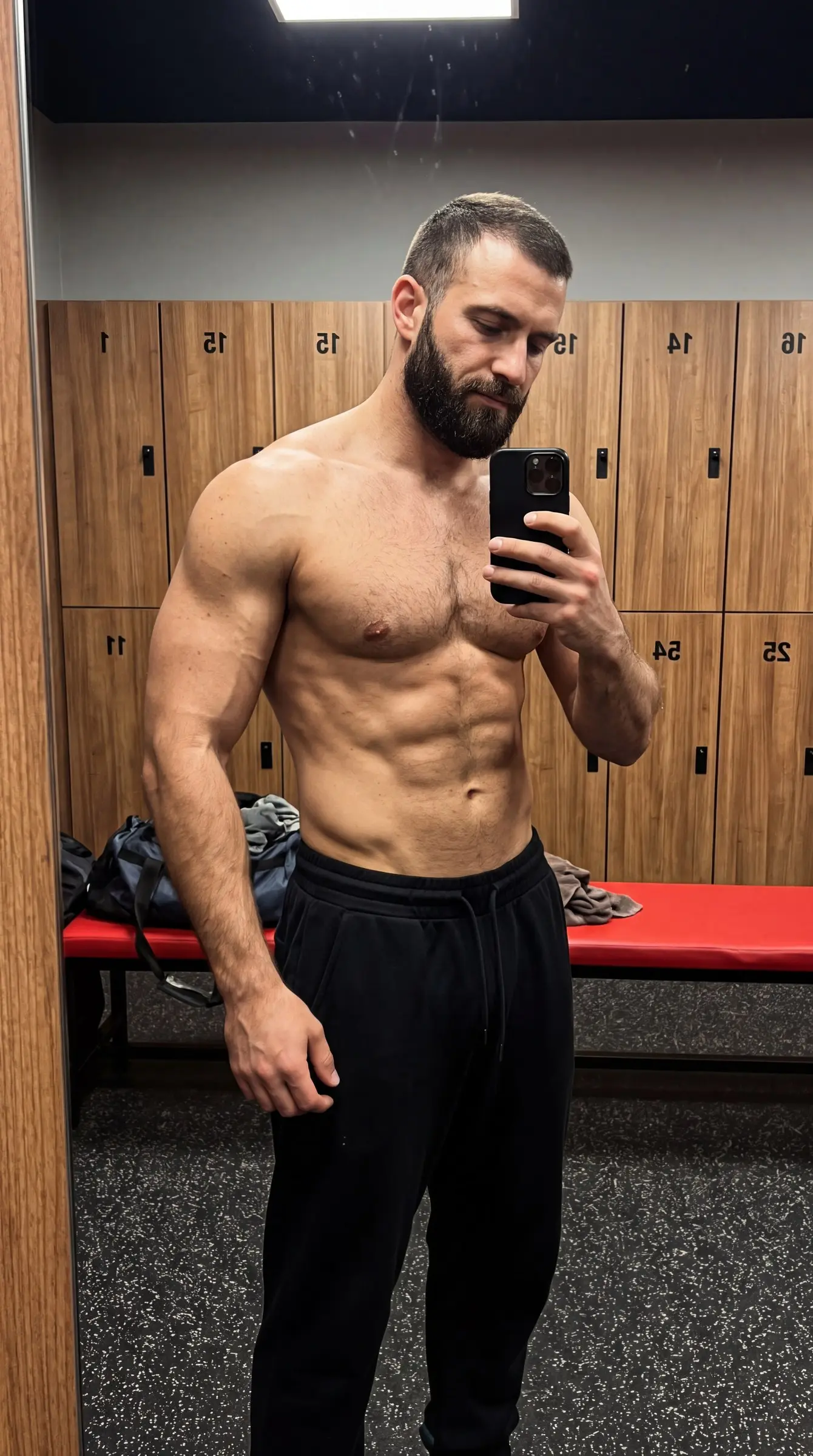 Shirtless bearded man in black sweatpants taking a phone selfie in a gym locker room with numbered wooden lockers and a red bench.