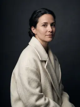 Woman with dark hair pulled back in an oversized ivory wool coat, three-quarter pose against a charcoal studio backdrop