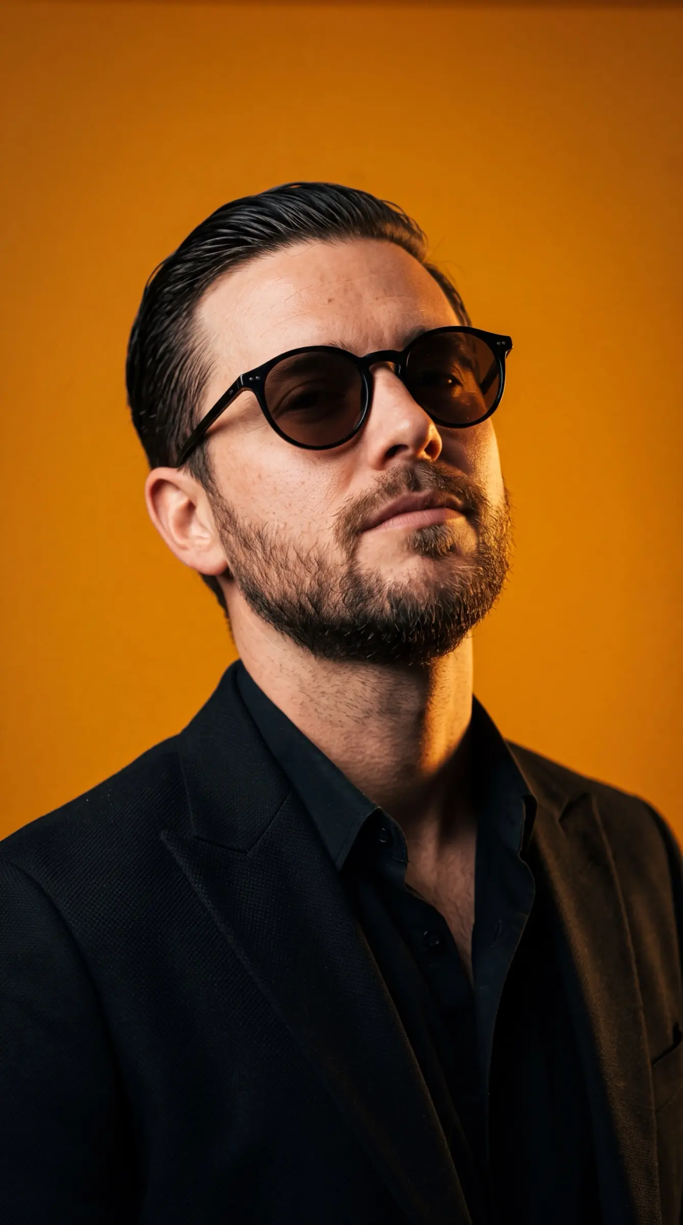Man in a black blazer and round sunglasses against an amber studio backdrop