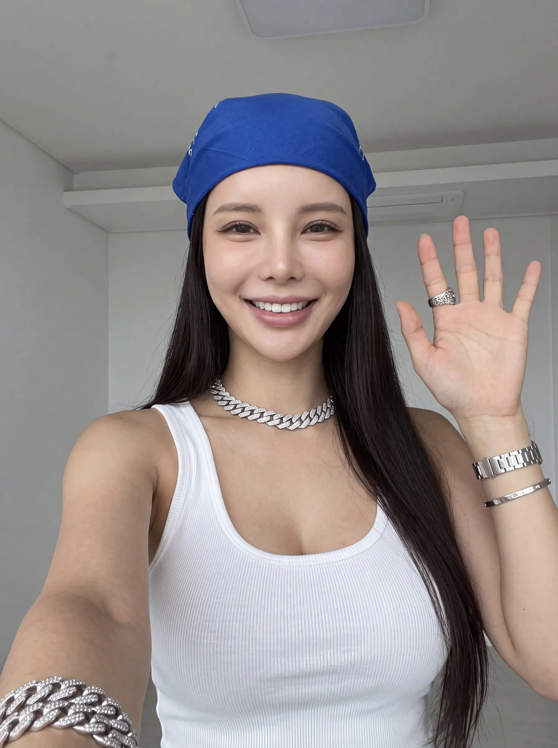 Smiling woman in a blue bandana waving in a close selfie against a white wall